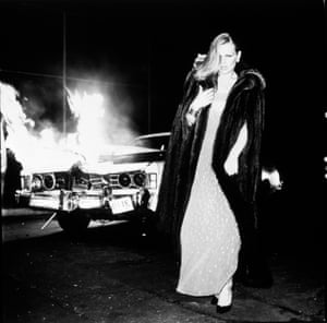 Patti Hansen, in finery of gown, fur coat and hair do, walks away from a burning car. This is glamour augmented with cinematic drama.