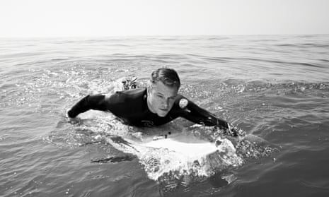Matt Damon in a wetsuit lying on a paddle board, paddling, in the sea
