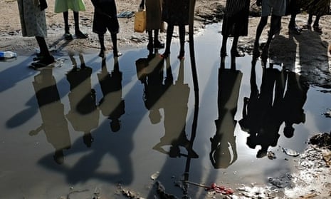 The shadows of Sudanese women and children are reflected in a pool of standing water as they wait at a water distribution point in the village of Guit.